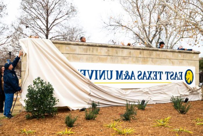 Several people stand behind the sign and release the canvas covering over the sign. A man on the far right reaches up to finish removing the canvas covering.